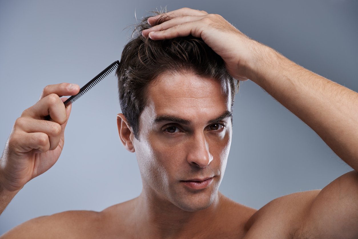 Mann stylt sich vor einem grauen Hintergrund mit einem Kamm seine braunen Haare und sieht in die Kamera